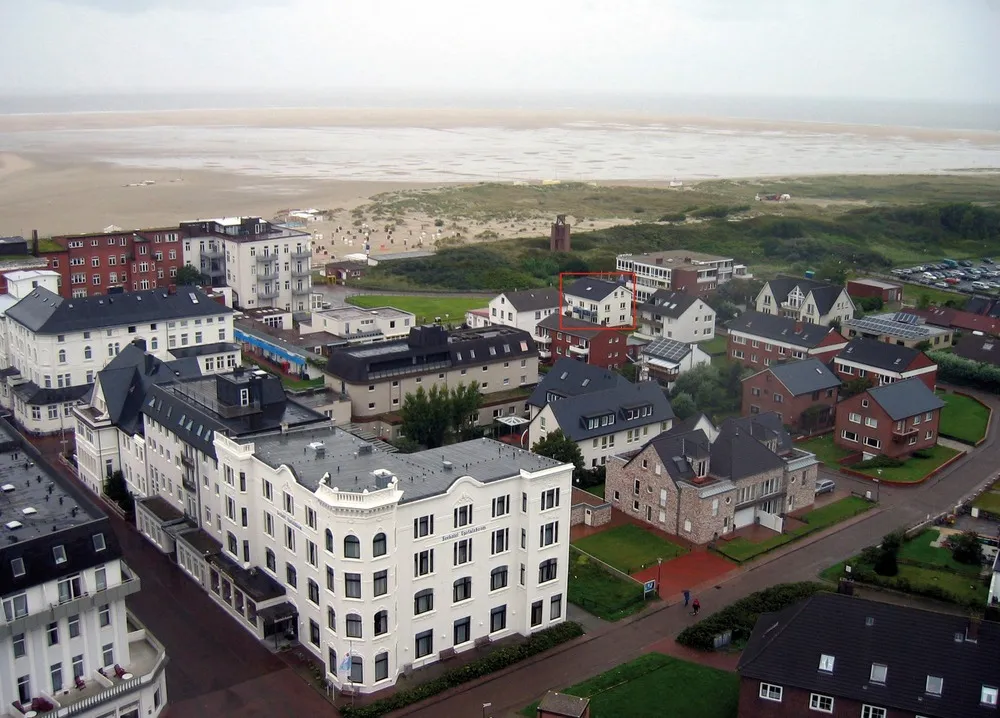 Luftaufnahme von Borkum mit dem rot markierten Haus Am Westkaap zwischen den Ortshäusern und dem grünen Dünenstreifen, dahinter der breite Sandstrand und das Meer.