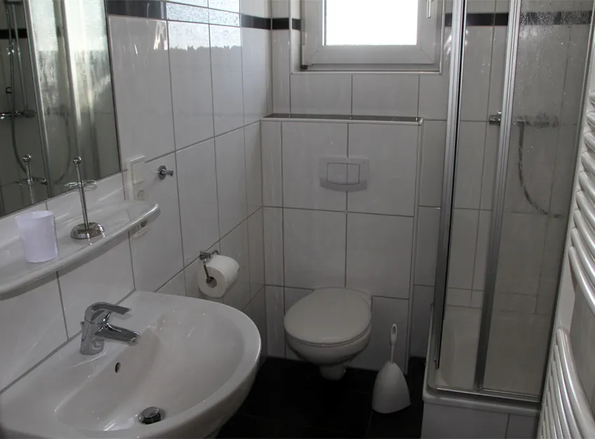 Weiß gefliestes Duschbad mit Waschbecken und Spiegel, Hänge-WC, Duschkabine mit Glastüren, Handtuchheizkörper und kleinem Fenster.