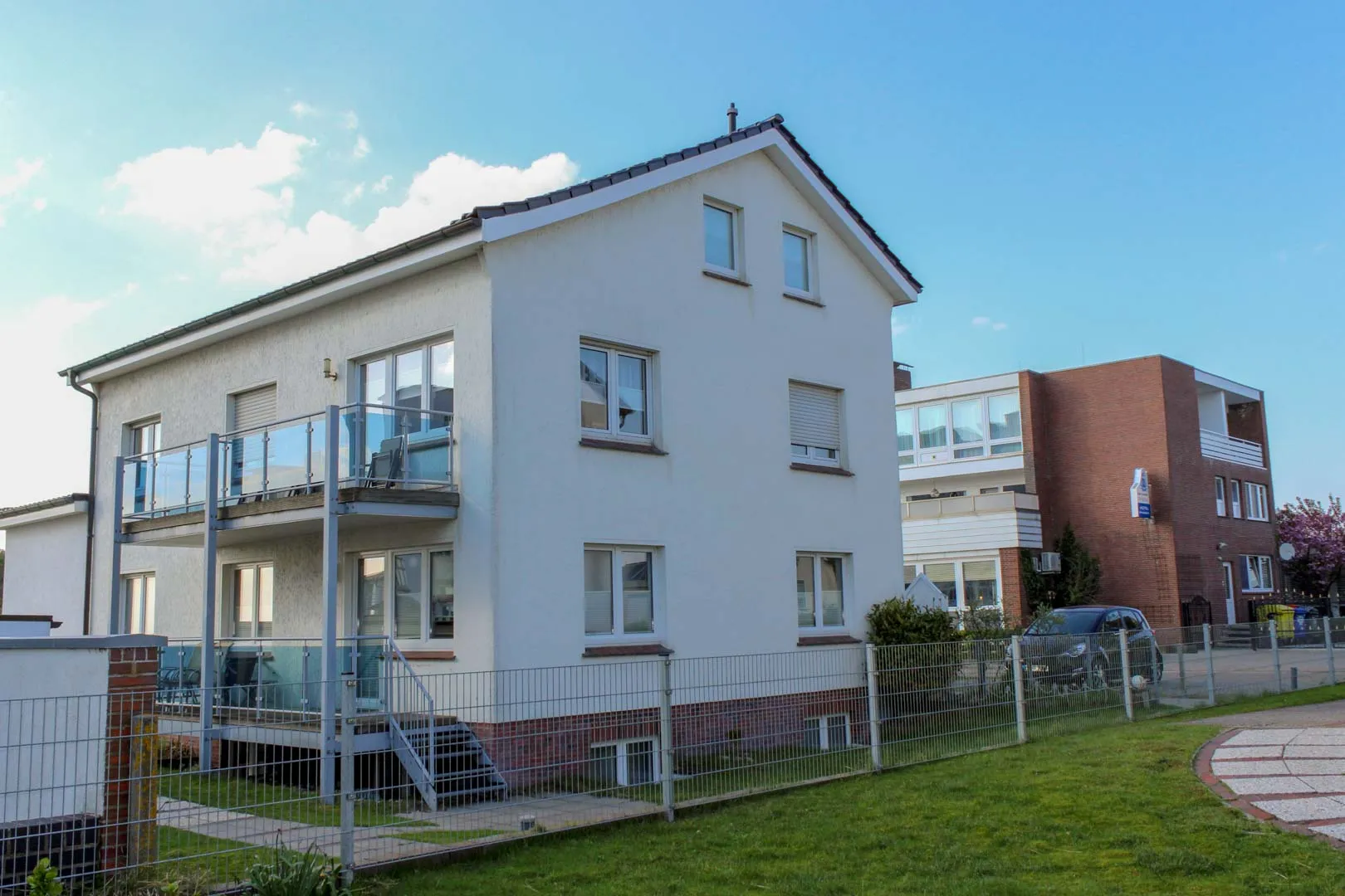 Weißes Haus Am Westkaap von außen mit umlaufendem Balkon, eigener Außentreppe zur Wohnung und eingezäuntem Garten unter blauem Himmel.