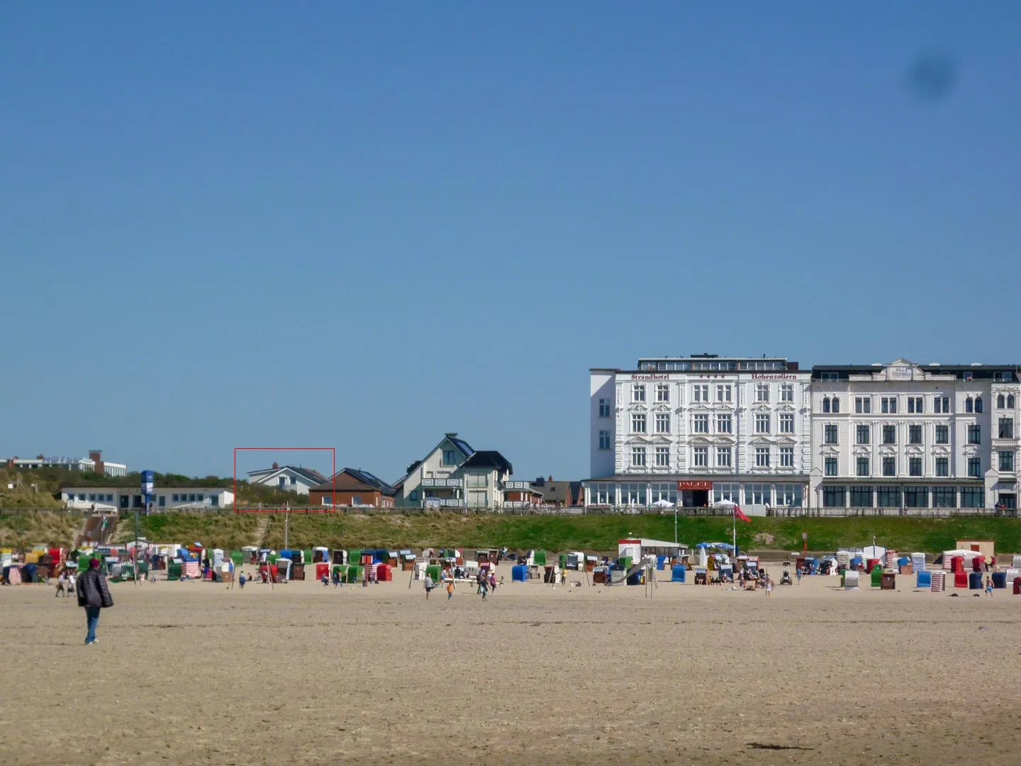 Nordstrand mit bunten Strandkörben und Spaziergängern, dahinter die Häuser an der Promenade, links das rot markierte Haus mit der Ferienwohnung.
