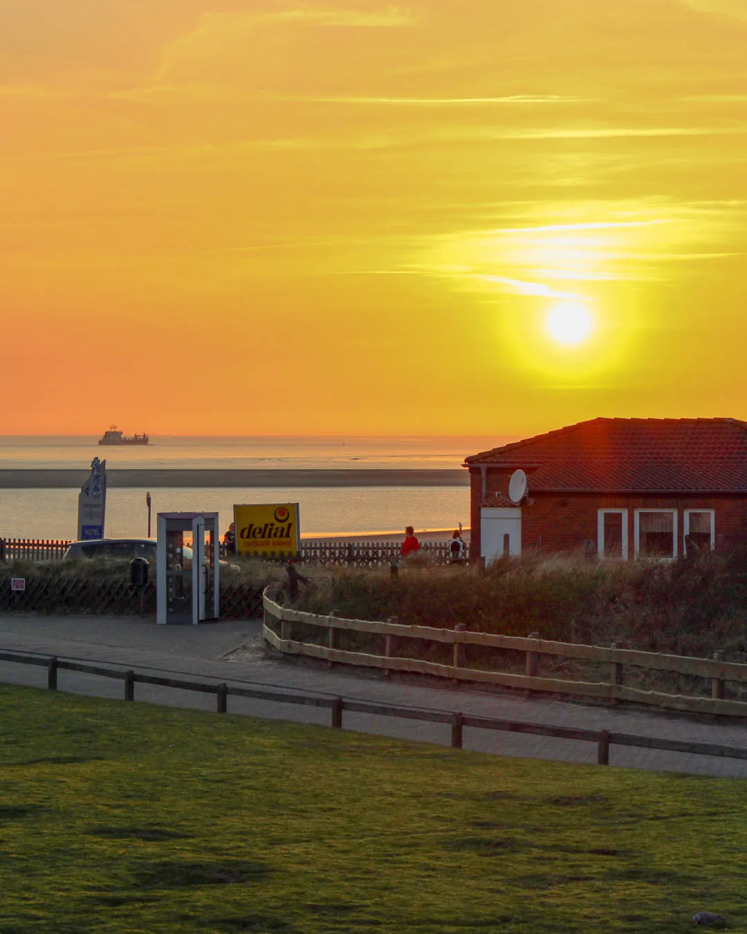 Sonnenuntergang in Orangetönen über der Nordsee am Strandaufgang neben dem Haus, mit rotem Backsteinhaus, Holzzäunen und einem Schiff am Horizont.