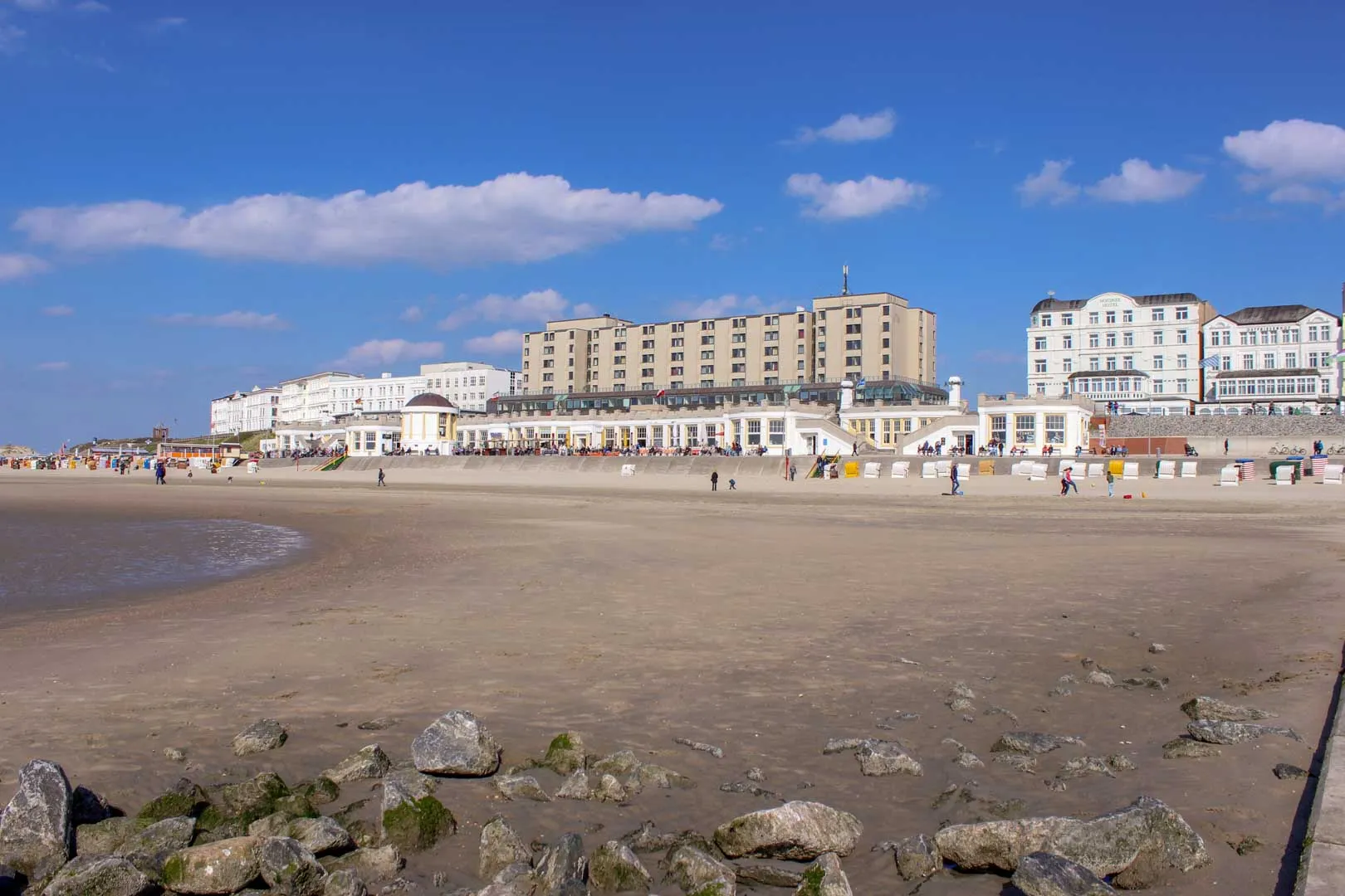 Weitläufiger Sandstrand bei Ebbe mit Blick auf die Wandelhalle und die Hotelfassaden an der Strandpromenade unter blauem Himmel.