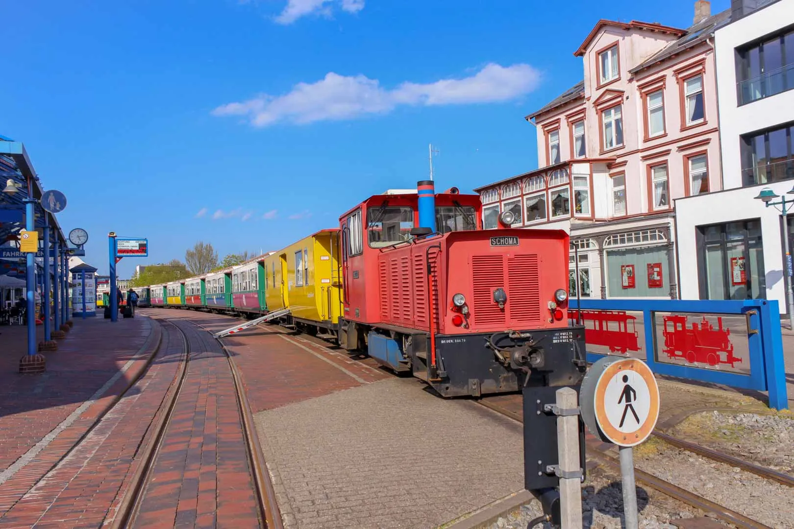 Die Borkumer Kleinbahn am Bahnsteig: eine rote Diesellok mit bunten Wagen vor den Häusern am Bahnhof.