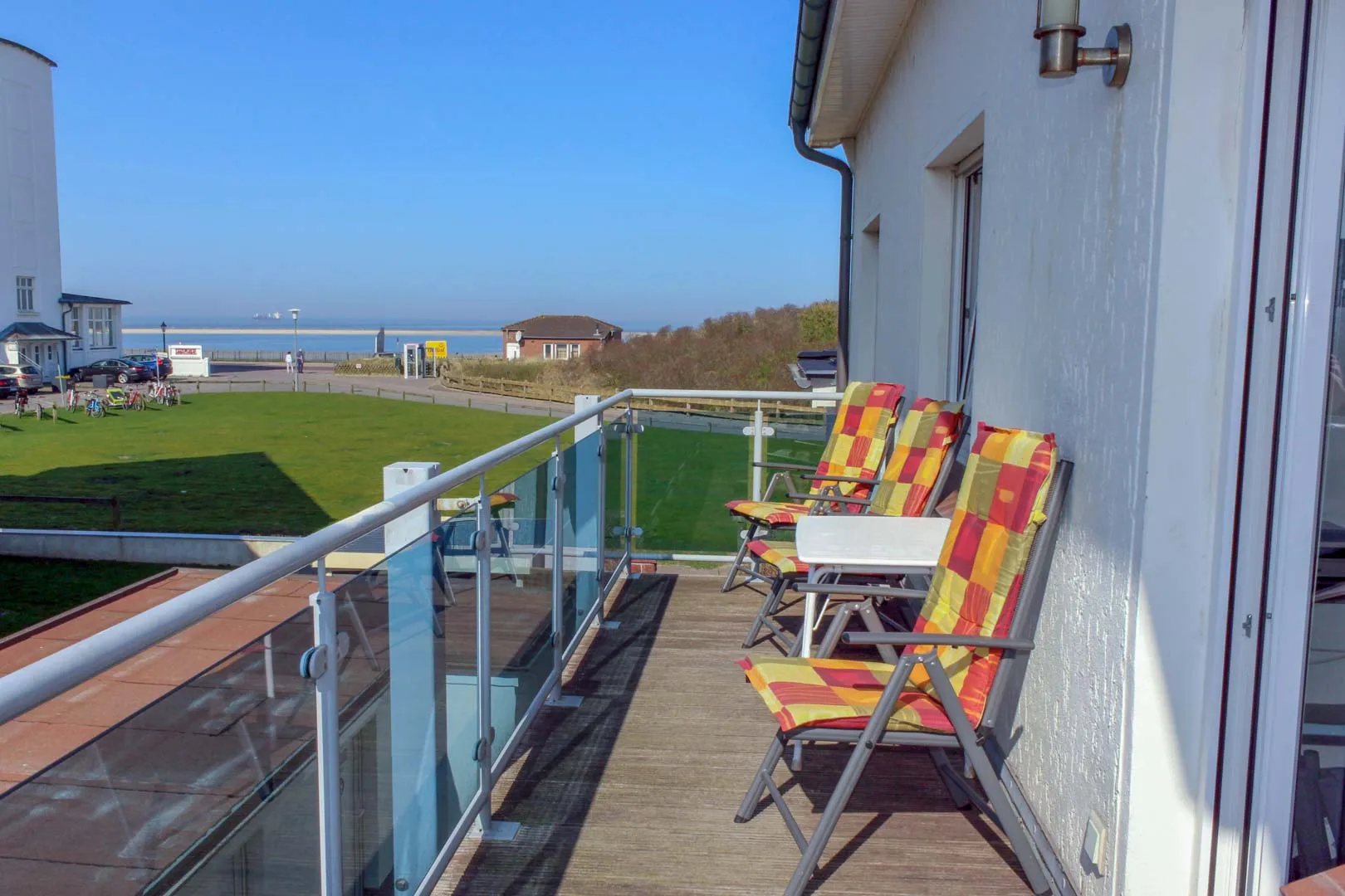 Sonniger Balkon mit Holzdielen, Glasbrüstung und drei Klappstühlen mit rot-gelb karierten Auflagen, dahinter Rasen, Promenade und die Nordsee unter blauem Himmel.