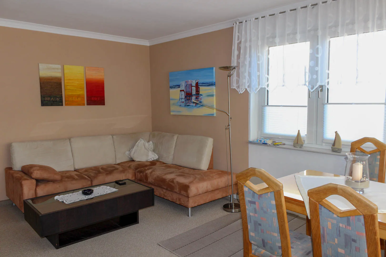 Wohnzimmer mit beiger Ecksofagarnitur, dunklem Couchtisch, Strandkorb-Gemälde und dreiteiligem Wandbild, rechts der Essplatz am hellen Fenster.