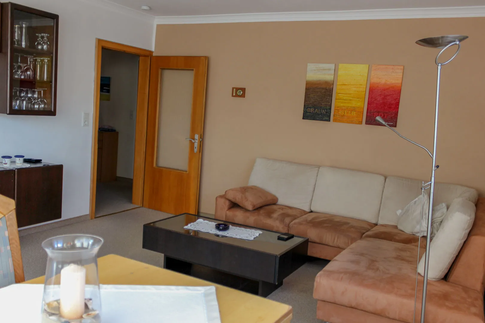 Blick durchs Wohnzimmer zur geöffneten Holztür in die Diele, links eine Glasvitrine mit Gläsern, rechts die Ecksofagarnitur unter drei farbigen Wandbildern.