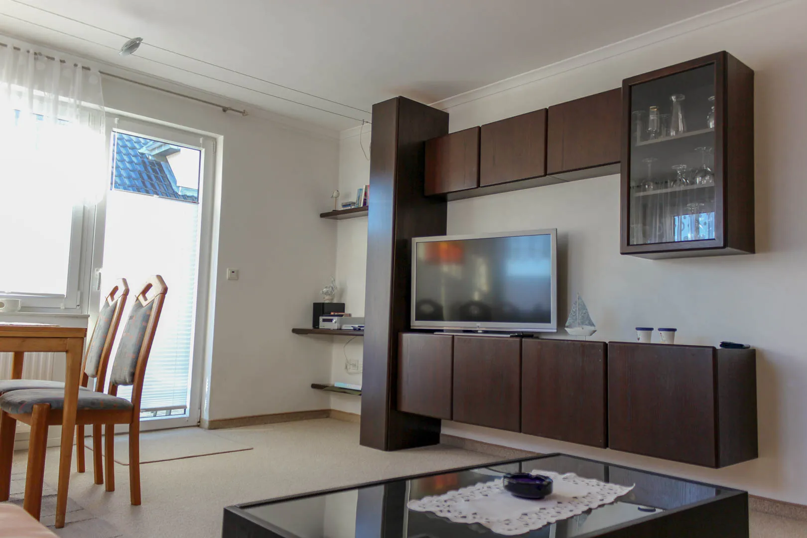 Dunkelbraune Wohnwand mit Flachbildfernseher und Glasvitrine im Wohnzimmer, links der Esstisch vor der bodentiefen Balkontür.
