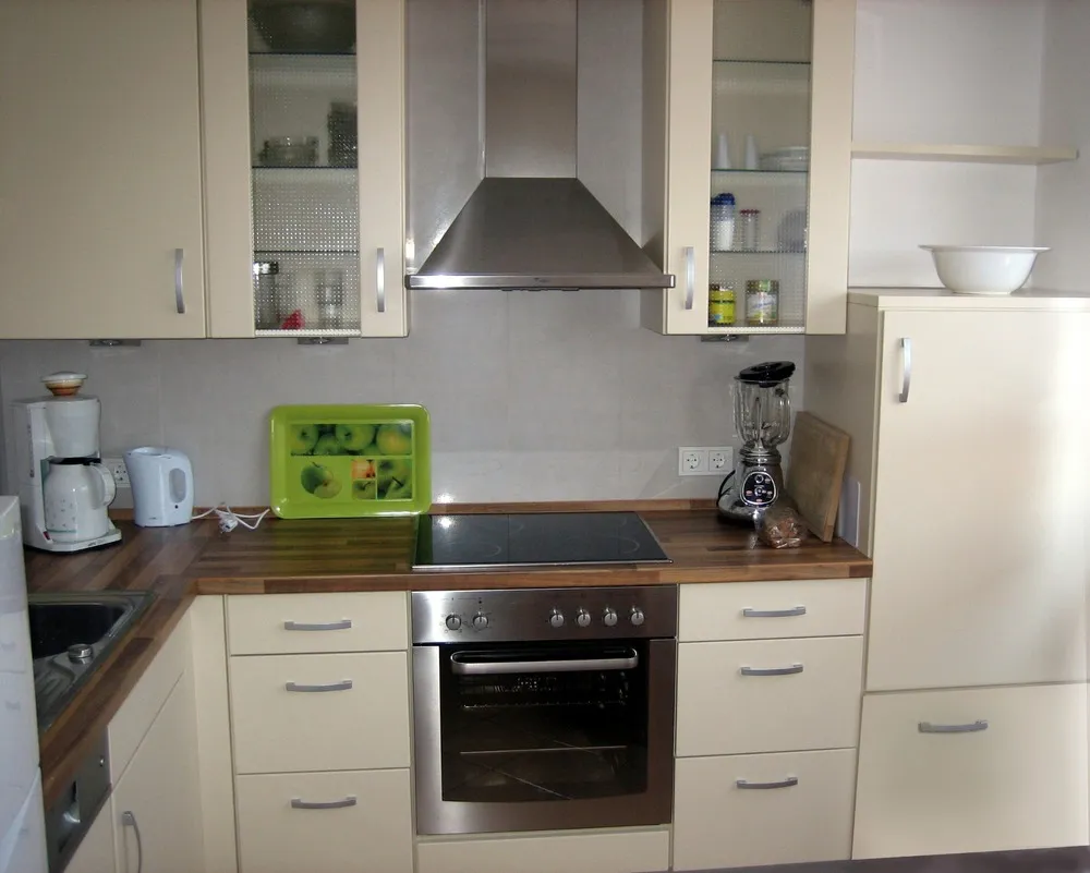 Küche mit cremefarbenen Fronten, Edelstahlbackofen und Dunstabzugshaube, Ceranfeld in der Holzarbeitsplatte sowie Kaffeemaschine, Wasserkocher, Mixer und Kühlschrank.