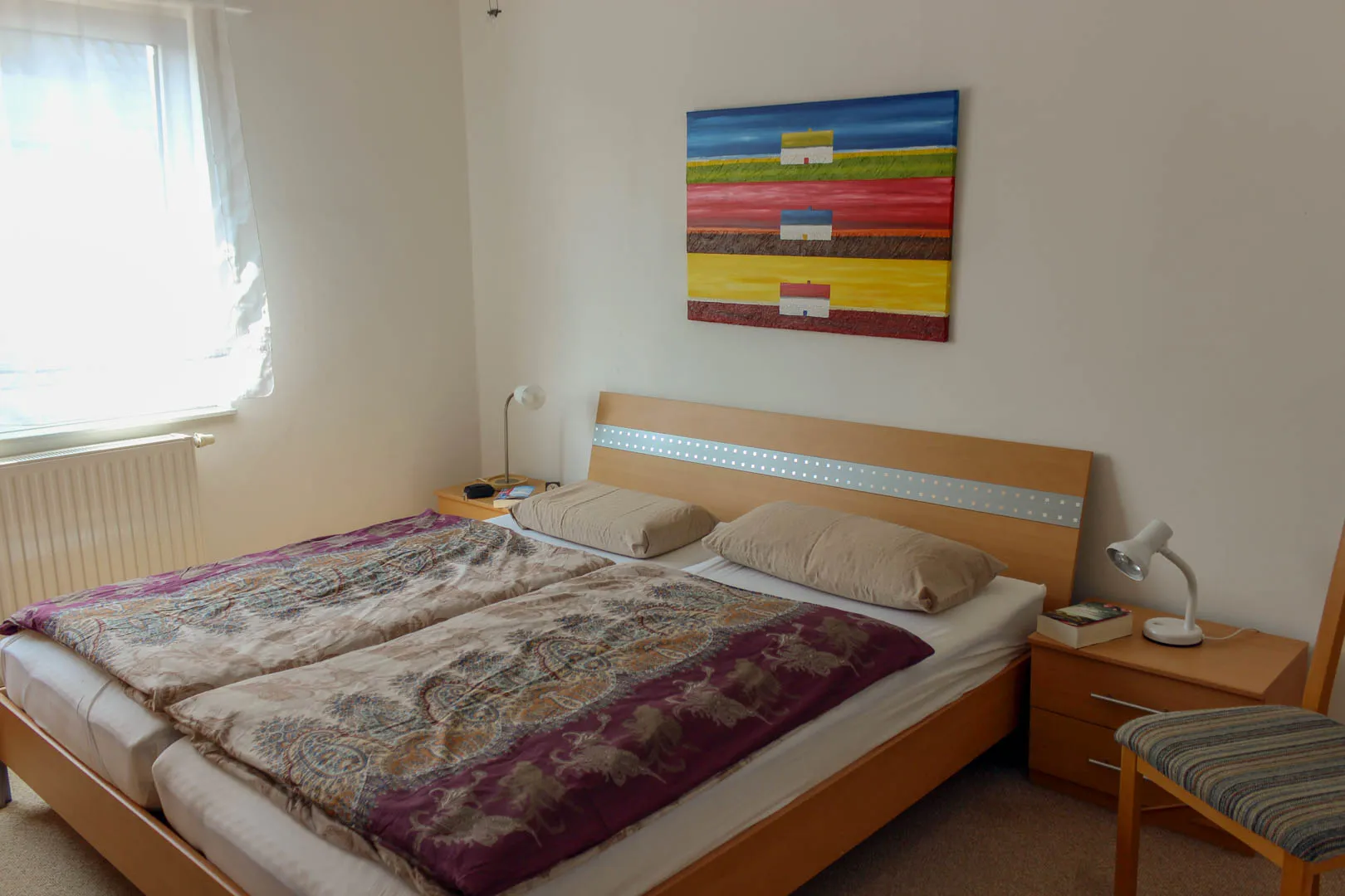 Schlafzimmer mit Doppelbett aus hellem Holz, zwei Nachttischen mit Leselampen und einem farbenfrohen Gemälde über dem Kopfteil, links das Fenster mit Heizung.