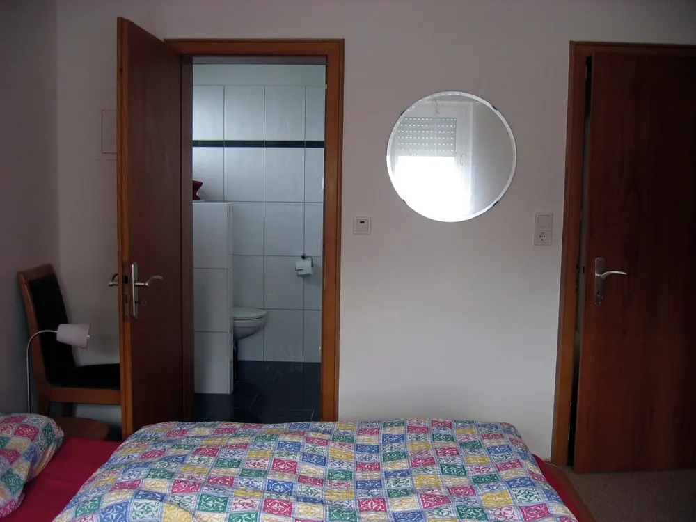 Schlafzimmer mit bunt gemustertem Bett und rundem Wandspiegel, durch die offene Tür Blick ins eigene weiß geflieste Duschbad mit WC.