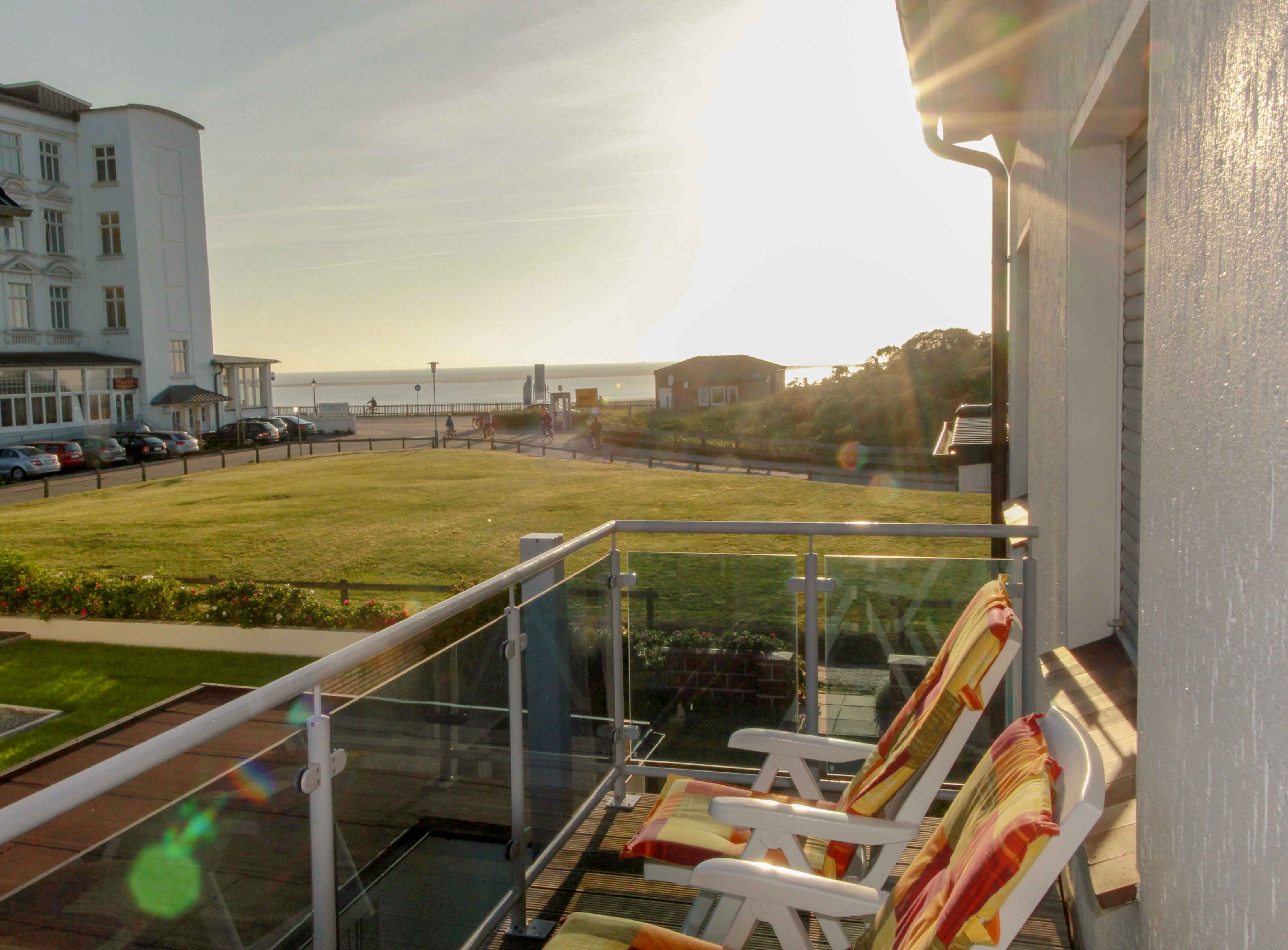 Abendlicher Blick vom Balkon über die Glasbrüstung und zwei Gartenliegen hinweg auf Rasen, Promenade und die Nordsee in der tiefstehenden Sonne.